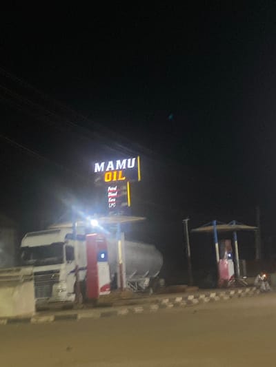 At night, the illuminated "MAMU OIL" sign advertises petrol, diesel, kerosene, and LPG in Unguwar Fatika, Nigeria. A tanker truck sits in the foreground. Other infrastructure is visible. The ambient light is low.