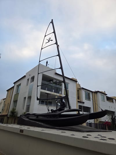 A bronze sculpture of a Hobie Cat sailboat with a figure seated in it is displayed in front of a modern, multi-story building complex in Dana Point, California. The sculpture is positioned on a concrete pedestal with numerous raised, dark grey, pebble-like indentations. A small black and gold plaque on the pedestal reads, "PLEASE KEEP OFF MONUMENT".

The sailboat sculpture is dark bronze, featuring two hulls, a mast with a stylized sail, and a seated figure appearing to steer the boat. The sail has a silhouette of a running figure within its frame.

Behind the sculpture, the building is white with beige and green accents, featuring balconies with glass railings. Windows and doorways are visible on the facade. The sky is overcast with diffused sunlight. To the right of the building, a dark fence and some bright pink flowers are visible.

The image is taken from a low angle, looking up at the sculpture and the building. The overall setting suggests an outdoor public space, possibly a waterfront area or a plaza.