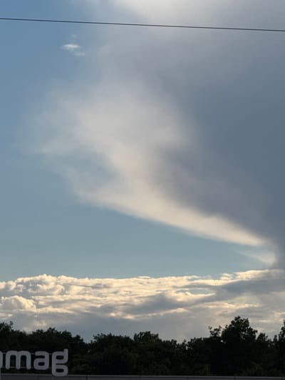 The image shows a vast expanse of sky with a variety of clouds. A thin, dark horizontal line, likely a wire or cable, bisects the upper portion of the frame. The sky transitions from a clear light blue on the left to a more overcast, gray hue on the right, with a large, wispy cloud formation dominating the center. Below this, a band of fluffy white clouds is visible. The bottom of the image features a dense line of dark green trees, likely forming a treeline or forest edge. In the lower left corner, a portion of a large white sign with the letters "mag" is visible, suggesting an outdoor advertisement or branding. The overall scene is an outdoor environment, likely a suburban or industrial area, given the presence of the sign and the treeline, with an emphasis on the sky.