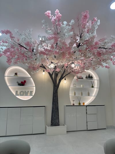 The image depicts an indoor scene, likely a commercial establishment, with a large artificial cherry blossom tree as the central focus. The tree has a dark brown trunk and branches adorned with pink and white blossoms. Several light bulbs hang from the branches, providing illumination.
On the left side of the tree, an oval-shaped wall niche with integrated lighting contains three shelves. The top shelf displays a small item, possibly a figurine or decorative object. The middle shelf holds three red candles and a small sprig of greenery. The bottom shelf features large white letters spelling out "LOVE."
To the right of the tree, a similar oval-shaped wall niche with lighting also has three shelves. The top shelf holds several small decorative items, including figurines that appear to be gnomes or similar characters, and miniature evergreen trees. The middle shelf contains a candle and several bottles, possibly lotions or diffusers. The bottom shelf displays reed diffusers and a small glass jar containing red beads or candies.
Below the niches and flanking the tree are two long, white cabinets with a minimalist design. The cabinets have a matte finish and appear to be made of wood or laminate. The cabinet on the left has doors and drawers, while the cabinet on the right has doors and drawers that are partially open, revealing contents.
The walls are painted a light color, and the ceiling is also light. The floor is tiled with light gray square tiles. A portion of a grey upholstered chair is visible in the bottom left corner of the image. The overall ambiance is clean, modern, and decorative.
The provided image does not contain any information about its location in Kettering, United Kingdom, nor does it depict any people, actions, or interactions.