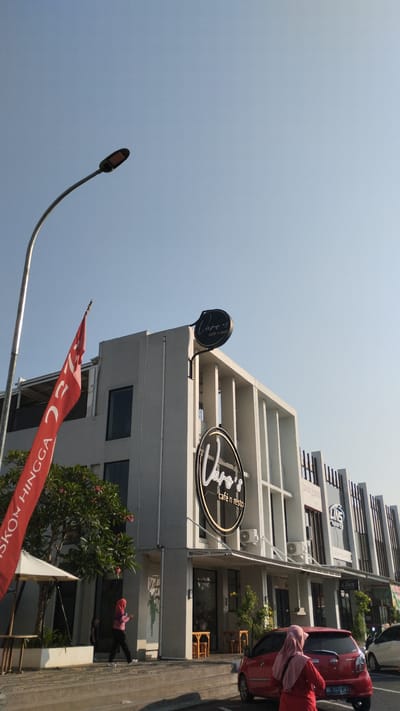 The image shows a street view in Trosobo, Indonesia, featuring a modern building housing a cafe and restaurant named "Varo's café n resto." The building has a light-colored facade with vertical architectural elements and several windows. A red flag with white text, partially visible, is planted in front of the building. A street lamp stands to the left, arching over the scene. Two women are visible: one wearing a pink hijab and black pants walks on the sidewalk, while another, also in a pink hijab and a red outfit, is near a red car parked on the street. The car's license plate is partially visible. Several small wooden tables and chairs are set up outside the cafe. The sky is clear and blue, indicating daytime. The overall scene depicts a commercial area with a focus on a cafe and its immediate surroundings.