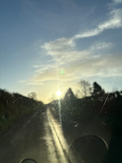 The image captures an exterior scene from inside a vehicle, viewed through a dirty or wet windshield, located in Ruyton-XI-Towns, United Kingdom. A prominent, low-lying sun is positioned near the center of the horizon, obscured and causing significant glare and lens flare, including a distinct green artifact above and to its left. Numerous illuminated dust particles, streaks, and potential water droplets are visible on the windshield surface, particularly in the lower half. A diagonal scratch or streak traverses the lower right quadrant of the windshield. The sky above is partly cloudy, displaying shades of blue, white, and light grey, with clouds illuminated by the low sun. The landscape, consisting of silhouetted trees and hills, is heavily blurred and appears as dark masses against the horizon. A road surface, highly reflective due to the sun's angle, extends forward from the vehicle, exhibiting two distinct parallel streaks of reflected light. No individuals or specific actions are discernable.