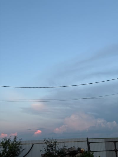 A low-angle, medium shot captures a sky with soft, pale blue hues gradually transitioning into gray clouds towards the horizon. Two distinct power lines stretch horizontally across the frame, creating a dividing line between the upper and lower portions of the image. Below the power lines, the sky is filled with scattered, fluffy clouds, some tinged with a soft pink or white. The bottom of the image features a white, corrugated metal fence running horizontally. On the left side of the fence, dark green foliage from trees and bushes partially obscures the fence. On the right side, the fence continues, with a dark metal gate or extension visible. There are no people or animals present in the scene. The overall environment appears to be an outdoor, possibly semi-rural or industrial, setting in Nizhniy Suyuk-Su, Russia, during twilight or early dawn, given the soft, diffused light and cloud coloration.