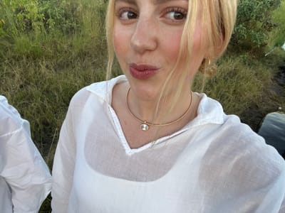 A young woman with fair skin and freckles, wearing a white sheer blouse over a white undershirt and a gold choker with a pendant, is taking a selfie. Her blonde hair is styled loosely around her face. Her expression is playful, with slightly pursed lips.

Behind her is tall grass, indicating a natural or semi-natural environment. The location is Lobilos, Botswana, suggesting the scene is likely outdoors in a rural or wilderness area. To the right, dark water is visible. A person wearing a white shirt is partially visible to the left of the subject.