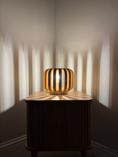 An indoor scene, captured at what appears to be night or in a dimly lit room, features a modern table lamp and a matching wooden cabinet or bedside table. The image is oriented sideways, but if rotated clockwise, the lamp stands upright on the table.

The lamp has a distinctive design with horizontal wooden slats forming its shade, allowing light to emanate outward. It is brightly lit, with the warm-toned bulb visible inside, casting strong, elongated horizontal striped shadows and light patterns across a light-colored wall to its left. The table, a rectangular piece with rounded corners, shares a similar slatted wooden design on its sides. The top surface of the table, directly beneath the lamp, reflects radial patterns of light and shadow, highlighting the lamp's base. The rest of the room, including the wall to the right and floor/ceiling areas, is cast in deep shadow, emphasizing the lamp's illumination.

The overall atmosphere is warm and inviting, showcasing contemporary design and dramatic lighting effects. There are no people visible, nor is there any discernible text. The location context for this image is Harrogate, United Kingdom.