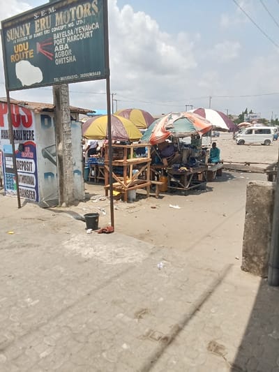 The image shows a street scene in Ovwian, Nigeria, located at coordinates 5.496 latitude and 5.797 longitude. The scene appears to be in front of a business called "Sunny Eru Motors," which is a division of Sunny Eru Concept Nig. Ltd. The business services the following areas: Bayelsa/Yenagca, Port Harcourt, Aba, Agbor, Onitsha. There are several small businesses selling items under umbrellas on the side of the road. There is one person visible, wearing a light-colored shirt and dark pants, standing behind a stall under an umbrella. Several objects can be seen, including a black bucket, a small table with a black cloth, and a sign advertising services. The scene is mostly paved. The weather appears to be sunny and dry.  The scene suggests that this is an everyday sight in Ovwian.  The street is narrow and appears to be a busy area.
