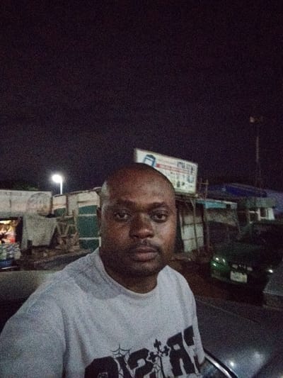 An adult male of dark complexion, with a shaved head and a short beard, is positioned in the foreground, gazing directly at the camera. He wears a light grey or white short-sleeved t-shirt featuring a dark, partially visible graphic design on the chest. The scene is set at night in Pasere, Nigeria. In the background, a bright, singular street light illuminates several rudimentary, single-story structures or market stalls. Light emanates from within some of these structures. To the right, a large signboard with a blue and white logo or text is affixed above additional background buildings. A dark-colored vehicle, possibly a sedan, is parked on the right side of the street, with its rear facing the viewer. The entire image exhibits significant digital noise, indicative of low-light conditions.