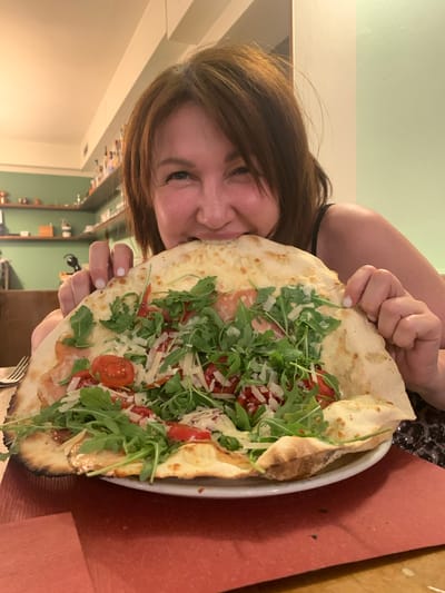 A woman with auburn hair smiles as she takes a bite from a large, thin-crust pizza topped with arugula, cherry tomatoes, and shaved parmesan cheese. She is holding the pizza with both hands, and her face is partially obscured by the food. The pizza is served on a white plate, which rests on a red placemat. In the background, shelves stocked with various items and a light green wall are visible, suggesting the image was taken in a restaurant in Montalcino, Italy.