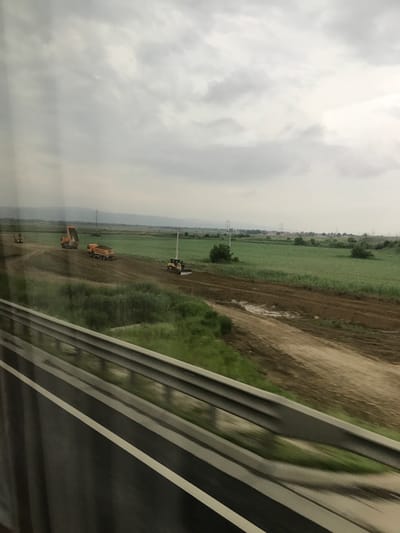 The image displays a construction scene in a rural or semi-rural landscape, possibly near Nicolae Bălcescu, Romania, viewed from a moving vehicle on an overcast day. In the immediate foreground, a grey metal guardrail runs diagonally from the lower left to the lower right, partially obscuring a dark paved surface with a white lane marker, both exhibiting motion blur. Beyond the guardrail, a strip of green vegetation is visible.

The middle ground is dominated by a wide, unpaved dirt construction path. From left to right on this path, a yellow excavator is partially visible, followed by an orange dump truck with its bed raised, likely in the process of unloading. A second orange dump truck, laden with earth, is positioned further right on the path. On the far right, a yellow bulldozer or grader is actively shaping the earth. The dirt path shows variations in color and moisture.

Beyond the construction activity, flat green fields extend into the background. Scattered utility poles with overhead lines are visible across the mid-distance. The horizon features faint, low-lying hills or mountains obscured by haze. The sky is uniformly overcast with light grey clouds, indicating diffuse lighting. No individuals are explicitly visible in the scene.