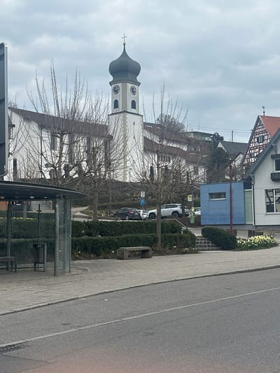 The image depicts a street scene in Krauchenwies, Germany, under an overcast sky. In the foreground, an asphalt road with a central lane marking is visible, bordered by a light-colored paved sidewalk. On the left side of the sidewalk stands a bus stop shelter constructed of glass and dark metal, containing a bench and a public waste bin. To the right of the bus stop, a stone bench rests on the sidewalk.
Beyond the sidewalk, a landscaped area features low, trimmed hedges, several bare deciduous trees with visible branches, and patches of yellow flowering plants, likely daffodils. An elevated paved area or street in the midground displays multiple parked vehicles, including a dark sedan and a white SUV. A circular speed limit sign (partially obscured) and a blue parking sign are visible in this area.
To the right in the midground, a modern, light-blue building with a horizontal window and a glass door stands next to a traditional white and brown half-timbered house with a red tiled roof and a weather vane.
Dominating the background is a white church building featuring a tall bell tower with a distinct dark, bulbous onion dome surmounted by a golden cross. The tower displays two clock faces, each showing the time as approximately 10:10 or 10:15. Other white buildings with brown roofs are visible behind and adjacent to the church. No individuals are present in the scene, and no specific actions or interactions are occurring.