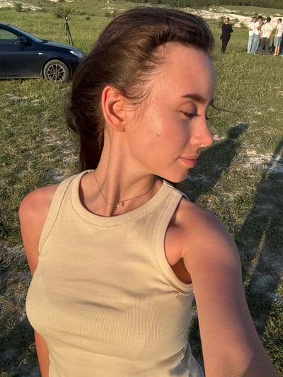 The image captures a young woman lying on grass near Вишенское сельское поселение, Ukraine. She is wearing a beige tank top and a delicate necklace. Her eyes are closed and she is facing upwards. A car is parked nearby, and in the distance, a group of people are visible. The scene appears to be outdoors in a grassy area under natural light.