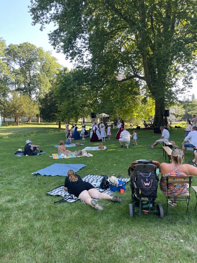 The media file depicts an outdoor gathering of people in a park-like setting, likely during the daytime, with sunny weather suggested by the lighting. Several individuals are seated or lying on blankets on a grassy lawn, facing a group of people who appear to be performers dressed in historical attire. Some of the performers are standing, while others are in poses suggesting a performance or reenactment. A large tree with dense foliage dominates the left side of the frame, casting shadows on the grass.
In the foreground, several objects are visible, including a stroller with a black bag attached, picnic blankets, hats, and shoes. One person in the foreground is lying on a blanket, facing away from the camera. Another person is seated, viewed from behind, wearing a colorful patterned dress. Further back, a group of people are observing the performance, with some seated on chairs and others on the ground. A speaker system and musical instruments are visible near the performers, indicating a possible concert or theatrical event. The overall atmosphere appears relaxed and leisurely, with people enjoying an outdoor event. The location context suggests this event is taking place in Stupava, Slovakia.