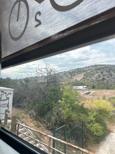 This is a landscape image taken from inside a vehicle, likely a bus or train, as suggested by the window frame and textured screen in the foreground. The image is oriented vertically.
The scene depicts a mountainous area with a road running through it. A white truck with a blue stripe is visible on the road, approaching a tunnel entrance carved into the hillside. The mountain is covered in sparse vegetation, with rocky outcrops. To the right of the road, there is a dense patch of green trees and shrubs, with dry grass and a metal fence visible further down. A rusty metal railing and stairs leading downwards are also present on the right side of the frame.
In the foreground, a large, partially obscured circular shape with a horizontal line inside it, and a letter "S" are visible, likely a logo or branding on the window screen. The sky is partly cloudy, suggesting it is daytime. The overall impression is one of travel through a rural or natural landscape. No people are clearly visible in the image. The graffiti on the white structure in the bottom right is illegible.
The location context provided is Salses-le-Château, France.