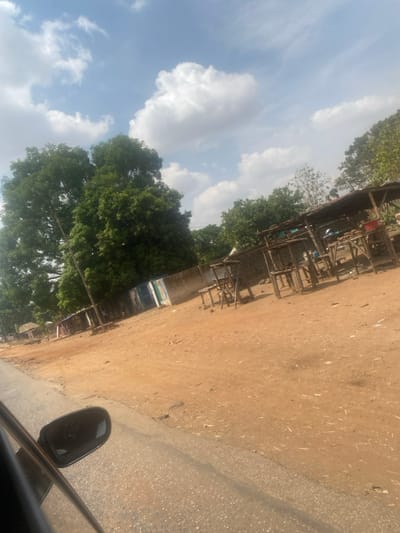 This image depicts a roadside scene in Tataura, Nigeria, under a partly cloudy blue sky. The foreground on the left shows a portion of a paved road and the black side mirror of a vehicle, from which the photograph was taken. The immediate right of the road is a wide expanse of reddish-brown dirt or laterite soil, which constitutes the ground for the rest of the scene.
On the left side of the dirt area, large, verdant trees dominate the background, with a slender pole vertically positioned near them. Adjacent to the trees and pole, low-rise structures with corrugated metal sections, some painted light blue and white, are visible behind a light-colored concrete wall running horizontally.
On the right side of the dirt area, a series of rustic, open-air market stalls or stands are present. These structures feature simple wooden frames and shaded roofs, appearing largely unoccupied. One stall on the far right contains a red basin and what appear to be agricultural produce or goods. No individuals are visible in the scene. The overall environment suggests a semi-urban or village setting with commercial activity.