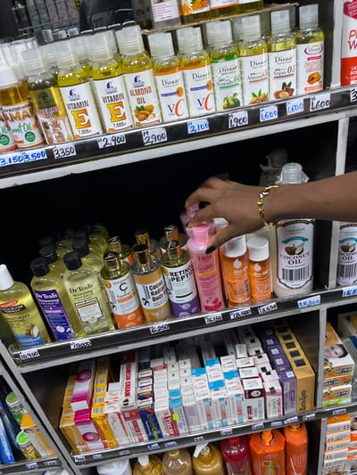 An interior retail scene in Alasia, Nigeria, depicts three shelves stocked with various personal care products. On the top shelf, numerous small bottles of cosmetic oils such as Vitamin E, Almond, Avocado, Argan, and Papaya are displayed, with prices ranging from ₦1,600 to ₦3,500. The middle shelf features larger bottles of bath and body oils, lotions, and serums, including Dr. Teals, Palmer's Cocoa Butter, Vitamin C serum, Cocoa Radiant Body Oil, Retinol Peptide, F&W Glycer Oil, Bio-Oil, and Orchedeuwa Organics Coconut Oil, priced between ₦6,250 and ₦25,300. A right hand with dark nail polish and a gold-colored heart-link bracelet is visible, reaching for a pink bottle of F&W Glycer Oil on this shelf. The bottom shelf holds stacked boxes of various creams and ointments, including "Roncin Cream" and "Temposat." All products are arranged on dark-colored shelves with white price tags indicating values in Nigerian Naira (₦).