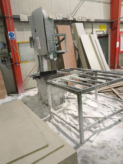In the Municipal Unit of Inofyta, Greece, a workshop scene is captured, dominated by a large, industrial band saw machine. The machine, covered in white debris, is positioned in the center of the frame, with its cutting blade visible. In front of the saw is a metal workbench, also heavily dusted with white material. The floor surrounding the machinery is littered with white scraps and dust, suggesting recent cutting activity. To the right of the saw, stacked panels of wood or similar material lean against each other. In the background, a red-painted support beam stands vertically. Further back, an illuminated exit sign in yellow with the word "Exit" and Greek text "ΕΞΟΔΟΣ" is visible above a doorway. The environment appears to be an indoor industrial setting with a light-colored wall and ceiling.