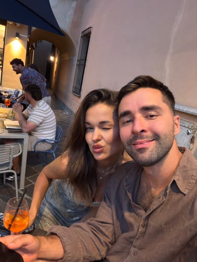 In Nobiallo, Italy, a couple is taking a selfie at an outdoor restaurant. The woman, on the left, is winking at the camera with her lips puckered. She is wearing a shimmering, sleeveless dress and a delicate necklace. The man, on the right, is smiling broadly, looking directly at the camera. He is wearing a brown button-down shirt. In the background, other patrons are seated at tables, and an umbrella is visible overhead, suggesting an awning or portable shade. The setting appears to be a narrow street or alleyway, typical of Italian towns, with buildings on either side. A glass of orange-colored beverage with a straw is in the foreground on the table.
