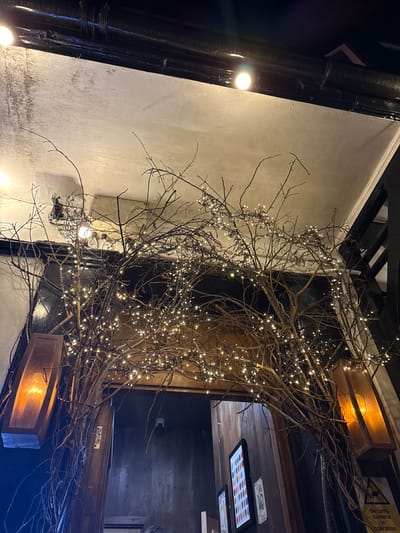 This appears to be an interior shot, possibly of a pub or restaurant in Windermere, UK. The image is taken from a low angle, looking up. A thick, black pipe runs vertically along the left side, with a few small lights attached. The wall is a pale, textured color. In the center is a display of bare branches adorned with small white lights, creating a visually prominent feature. This branch arrangement partially obscures a dark opening or doorway. Above the doorway, there's a wooden structure with what looks like signage. To the right of the doorway, one can see a glimpse into another room with a dark wooden wall, a few pictures, and a lighting fixture. The overall impression is of a cozy, dimly lit space with rustic decor.