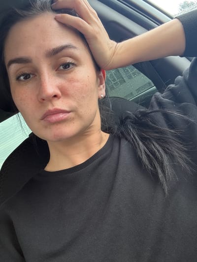 This media file is a close-up, low-angle portrait of a young woman, likely taken outdoors. She is resting her left arm on her forehead, with her hand splayed across her brow. Her eyes are dark brown, and her eyebrows are well-defined. She has a light complexion with visible freckles across her cheeks and nose. Her lips are full and appear to be slightly parted. She is wearing a black long-sleeved top. Her dark hair is loose and falls around her face.
The background suggests an outdoor setting, possibly inside a vehicle, with a window visible to the left. The window appears to have raindrops on it, indicating it might be raining or has recently rained. The lighting suggests it is daytime, but the weather might be overcast. The overall mood of the image is contemplative, perhaps a moment of reflection or fatigue. There is no visible text in the image. The provided location context is Verkhneyarkeyevo, Russia.
