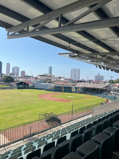 This image captures a wide-angle view of the Estadio Juan Demóstenes Arosemena baseball stadium, located in the Curundú district of Panama City, Panama. The perspective is from beneath the stadium's large, industrial-style metal roof structure, which features exposed steel beams and white undersurface panels with integrated light fixtures.
In the foreground and mid-ground, numerous rows of empty, dark green stadium seats are visible, extending along the first base line and into the lower right portion of the frame. The stadium's baseball field occupies the central and left areas of the image, showcasing a well-maintained green grass outfield and infield, delineated by reddish-brown dirt paths for the bases and a pitcher's mound. A black metal batting cage is positioned near the outfield fence, close to the first base line.
One individual, an adult male, is observed standing against a concrete barrier on the right side of the frame, partially obscured by the railing. He is facing away from the field and appears to be looking down at a handheld device.
Beyond the outfield, a dense urban environment stretches to the horizon. This includes a mix of lower-rise commercial and residential buildings, interspersed with green areas and several prominent high-rise skyscrapers forming a distinct city skyline in the distant background. Additional green stadium seating and various building structures are visible in the outfield perimeter. The scene is illuminated by strong sunlight under a clear, bright blue sky.