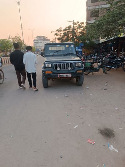 The scene depicts an urban street in Anantapur, Andhra Pradesh, India, under the soft light of dusk or dawn, characterized by an orange and pink sky.

In the mid-ground, a dark green or black Mahindra Bolero (likely a Camper variant due to its structure, though a full view of the rear is obscured) is parked facing the camera on the left side of the paved road. The vehicle's license plate, "AP21 BX 1254," confirms its registration in Anantapur district. A "5T" sticker is visible on the upper left of its windshield. The front bumper shows minor wear.

Two male individuals are walking away from the camera on the paved road, positioned to the left of the parked SUV. The individual on the left wears a black long-sleeved shirt and brown pants with white sneakers. The individual on the right wears a white long-sleeved shirt and dark pants with dark shoes. Part of a bicycle wheel is visible on the far left foreground.

To the right of the parked SUV, beyond the paved road, a dirt/gravel shoulder is present, heavily littered with paper, plastic wrappers, and other debris, including a dark, irregular stain near the SUV's right front wheel. Numerous motorcycles and scooters are parked along this shoulder, adjacent to a series of market stalls or shops with awnings and visible merchandise. One prominent building in the background on the far right appears to be an unfinished or under-construction multi-story brick and concrete structure. Further down the road on the left, another multi-story white building with a rooftop sign partially reading "Sree" is visible. Tall streetlights and power poles are distributed throughout the background, with various trees featuring green foliage.
