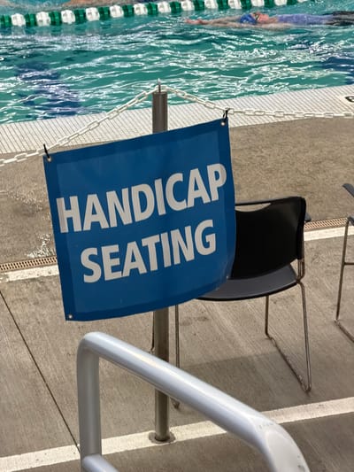 A blue banner with white lettering that reads "HANDICAP SEATING" is hanging from a metal pole in front of a black chair. The banner is positioned near the edge of a swimming pool, indicated by the presence of lane lines and a swimmer in the background. The ground around the pool is a light-colored concrete surface, and there is a white railing in the foreground. The scene appears to be an indoor swimming facility.