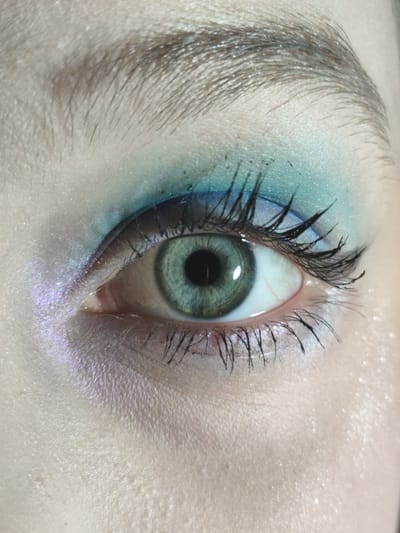 A close-up photograph of a human eye, specifically the left eye. The iris is green, with a dark pupil in the center. The sclera (white part) is visible, with some slight redness from blood vessels. The eye has been made up with blue and white eyeshadow on the upper eyelid. The eyebrows are brown. The surrounding skin is fair. The location cannot be determined from the provided image.