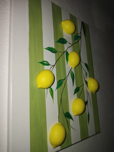A painting on a canvas features a striped background of white and lime green vertical stripes. The painting depicts a branch with several bright yellow lemons and green leaves. The lemons appear to be three-dimensional, possibly clay or another textured material, protruding from the canvas. The art is displayed on a wall. No specific location in Reftinsky, Russia can be determined from the image.