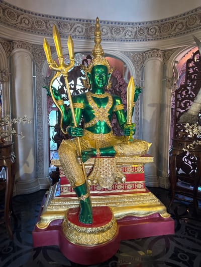 A vertically-oriented photograph features a green and gold statue seated atop a decorated pedestal. The statue, with its green skin, is adorned with gold embellishments and holds a golden trident in one hand and a golden sword in the other. It is seated on a raised, red and gold decorated throne. The statue is prominently displayed in a room with ornate white walls, with detailed carvings and floral designs. The floor appears to be tiled with dark geometric patterns. The location is the city of Samrong Nuea Subdistrict Municipality, Thailand.