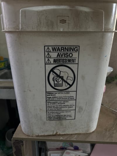 The image shows a white plastic bucket with a black warning label on it. The label reads "WARNING, AVISO, AVERTISSEMENT" and depicts an image of a child falling headfirst into a bucket of water. The label also includes a warning in English, Spanish, and French about the dangers of children falling into buckets and drowning. The bucket is sitting on a shelf. The location, according to the coordinates provided, is in the city of Pozos, Costa Rica. There are no people in the image.