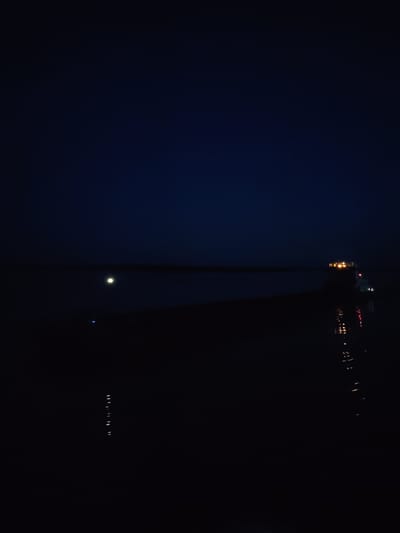A nighttime photograph shows a dark, calm body of water with a faint horizon line. A single bright light, possibly the moon, is visible in the sky. On the right side of the frame, a brightly lit vessel, likely a ship or ferry, is present on the water, its lights casting reflections. The overall atmosphere is serene and dark, with the artificial lights of the vessel being the most prominent feature against the natural darkness. The location is identified as Kudryashovo, Russia.
