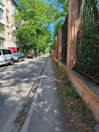 This image depicts a street scene in Sokolivske, Ukraine. The perspective is from a sidewalk, looking down a street lined with parked cars and a tall, weathered fence.
On the left side of the street, a multi-story apartment building is visible, its beige facade punctuated by numerous windows. Some windows have air conditioning units installed. Trees with lush green foliage cast dappled shadows across the street and sidewalk. Parked cars are a prominent feature, including a white van, a dark station wagon, and several other sedans.
To the right, a long, rust-colored fence with vertical black metal bars runs along the sidewalk. The fence is built on a low concrete base, and wild vegetation, including green leaves and dry brown debris, grows at its base and behind it. The fence and sidewalk are also shaded by the trees.
The sky above is a clear, bright blue, suggesting a sunny day. The presence of deep shadows indicates the time of day is likely mid-morning or early afternoon. The overall impression is one of a quiet residential street on a warm, sunny day. There is no visible text in the image.