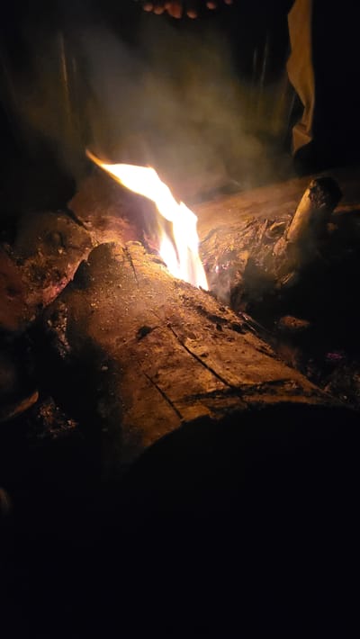 This image captures a vibrant campfire burning intensely at night, likely in Khir Ganga, India. The primary focus is on the large, rustic logs that make up the fire, with bright, yellow and orange flames erupting from the left side, casting a warm glow across the scene. Swirls of white smoke are visible, illuminated by the firelight as they rise.

The texture of the wooden logs, showing bark and splintered surfaces, is clearly discernible due to the strong light. In the lower-left corner, the bare toes of a person's foot are partially visible, suggesting someone is sitting very close to the fire, possibly enjoying its warmth or light. A piece of light-colored fabric, possibly clothing or a blanket, is also faintly visible in the upper-left background, catching some of the fire's glow.

The surrounding environment is cast in deep darkness, emphasizing the fire as the sole significant light source. The overall impression is one of warmth and comfort in an outdoor, nighttime setting, typical of a campfire experience in a natural environment like Khir Ganga. No visible text is present in the media file.