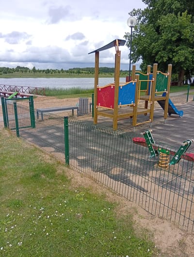 The image shows a children's playground located in Šaukėnai, Lithuania. The playground features a wooden play structure with colorful panels, a blue slide, and a seesaw with a spring mechanism. A green wire fence encloses the playground, separating it from a grassy area with small white flowers and a sandy path. In the background, a calm body of water, likely a lake, is visible, bordered by trees and vegetation. The sky is overcast with clouds. A lamppost stands near the play structure, and a trash can is situated on the playground's rubberized surface. A wooden bench is also present near the fence.