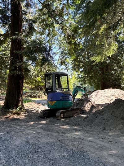 The image depicts a blue and green mini excavator situated on a gravelly area, likely a construction or landscaping site. To the left of the excavator is a large tree with dense green foliage, suggesting a forested area. The excavator appears to be working against a rock face or a large boulder. The setting is suggestive of the Bowen Island Municipality, Canada, given its natural landscape and likely construction needs. The excavator is the only object in the image. No people are present.