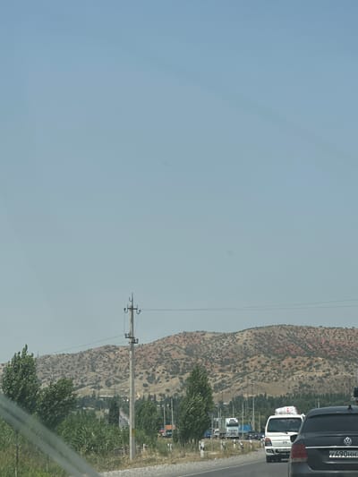 This is a photograph taken from a vehicle, showing a road with traffic in Kumush-Aziz, Kyrgyzstan. In the foreground, the right side of a black Volkswagen sedan is visible, facing away from the camera. Its rear license plate reads "Y490HH". To the left of the sedan, a white van with a white package strapped to its roof is also in traffic. Further down the road, a white truck with a visible license plate "670 ALC" is seen, along with other vehicles. The road is flanked by green vegetation and trees on both sides, with a power pole visible in the middle ground. In the background, a rugged, reddish-brown mountain range with sparse vegetation stretches across the horizon under a clear blue sky. The overall scene depicts a typical day on a highway with moderate traffic, set against a backdrop of natural landscape.