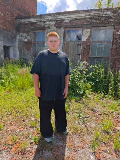 A young male with short red hair stands in the foreground, facing forward. He wears a loose-fitting black t-shirt and black pants. The background features an abandoned brick building with broken windows and peeling paint. The ground is covered in dirt, scattered bricks, and overgrown green grass and weeds. The sky above is blue with scattered white clouds. The scene appears to be outdoors, possibly in an industrial or derelict area within Cheremushki, Russia.