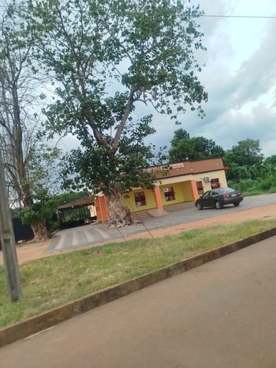 The image shows a one-story yellow and brown building with a sign that appears to say "GOOD". A dark-colored car is parked near the building. The building is located near the city of Abube-Nando in Nigeria. Large trees with green foliage frame the building. A covered area with chairs and tables is visible to the left of the building. There is grass and a paved area in front of the building. A road is visible in the foreground. The sky is cloudy. A utility pole is on the left side of the image.