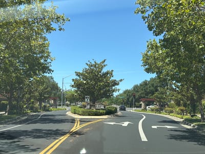 A driver's perspective shot captures a clear, sunny day in Elk Grove, California, with a focus on a road leading towards a roundabout. The road is asphalt, marked with yellow double lines on the left and white lane dividers. White arrows on the road indicate directions for traffic, with a left turn arrow prominently displayed in the foreground.
The roundabout itself is circular, featuring a central island with a manicured hedge and a decorative stone structure with a tree. The island is surrounded by a concrete curb. Beyond the roundabout, the road curves to the right.
Lush green trees with dense foliage line both sides of the road, with their branches extending into the bright blue sky. In the background, there are more trees, some residential-style buildings with stone accents and covered patios, and streetlights. A few cars are visible on the road in the distance, and a traffic light is illuminated red. The overall scene depicts a well-maintained suburban street.
