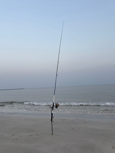 A fishing rod stands upright on a sandy beach in Ajman, United Arab Emirates. The rod, with a blue and white handle, is equipped with a fishing reel and is angled towards the calm sea. Gentle waves lap the shore, and a faint haze hangs in the sky, obscuring the horizon. In the distance, a long pier or breakwater stretches into the water, with a lighthouse or structure visible at its end. A ship is also discernible on the horizon. The beach is mostly empty, with only a few small shells and footprints scattered across the sand. The overall impression is one of peaceful solitude.
