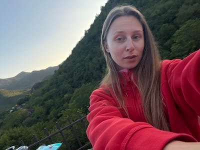 A selfie shot shows a young, fair-skinned woman with long, light brown hair wearing a red zip-up hoodie. She has a nose piercing on her left nostril. The background features a lush, green mountainside, with trees covering the slopes, partially illuminated by sunlight. In the distance, more mountains are visible under a pale sky. A black metal railing is seen in the foreground, with some objects, including what looks like a bag, partially visible below. The photograph appears to be taken in an outdoor setting, specifically, Dupilo, Montenegro.