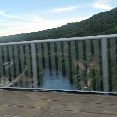 This is a high-angle, medium shot of a river valley with a forested hill in the background. The sky is blue with scattered clouds. A grey metal railing with vertical glass panels runs across the foreground, partially obscuring the view of the scene below. Through the glass panels, a river is visible, flowing through the valley. On the left side of the valley, a building with a light-colored facade and a dark roof can be seen, along with some paved pathways. The hill in the background is densely covered with green trees. The image is taken from a bridge or an elevated viewpoint.