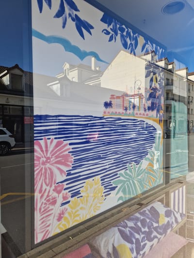 A reflection in a shop window on a street in Antony, France, displays a colorful mural. The mural depicts a stylized beach scene with blue striped water, a sandy shore, and a row of colorful buildings with palm trees. In the foreground of the mural, there are abstract floral shapes in pink, yellow, and green. The reflection also shows buildings across the street, a white car, and people. Below the mural, inside the shop, several pillows with floral patterns are visible. The street outside appears to be paved, with a yellow line marking a lane.