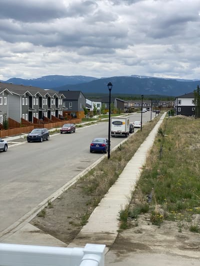 This image depicts a residential street in Whitehorse, Canada, under a cloudy sky. The street features a paved roadway with sidewalks on both sides. Houses, primarily townhouses and single-family dwellings with modern architectural styles and neutral color palettes, line the left side of the street. A few cars are parked on the street or in driveways. A white delivery truck with "Certified Heating & Service" branding and a phone number is parked on the right side of the road. In the background, forested hills and distant mountains, some with patches of snow, are visible. The right side of the street is a grassy slope with some yellow wildflowers and sparse vegetation. A white railing is visible in the foreground, suggesting the photo was taken from a porch or balcony.