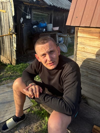 A man sits on a wooden bench in front of a dilapidated wooden structure, likely in Sovkhoza "Razdol'e", Russia. The structure has a corrugated metal roof and appears to be in disrepair, with peeling paint and exposed wood. The man, with a buzz cut and blue eyes, wears a black sweatshirt and shorts. His hands are clasped, and he has tattoos on his left forearm. A ring is visible on his left ring finger. He is wearing a sandal with a black sock. The ground is covered in dirt and sparse vegetation. In the background, there are various objects, including plastic crates, a bucket, and what appears to be a metal basin. The scene is illuminated by sunlight, casting shadows.