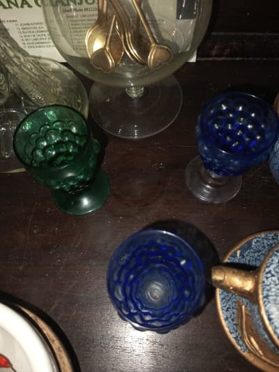 The image showcases a collection of glass objects on a dark surface. A large glass container filled with spoons with heart-shaped details is prominent. Beside it, a green glass cup with a textured surface sits. Two smaller blue glass cups, also with a textured pattern, are positioned nearby. Finally, a teacup with a blue and white pattern and a golden handle rests beside one of the blue cups. The composition is tightly cropped, focusing on the arrangement of these glassware pieces.