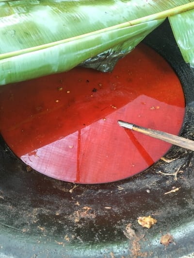 A close-up, top-down perspective captures the interior of a large, dark, circular cooking vessel located in Jambo Dalam, Indonesia. The vessel holds a viscous, reddish-orange liquid, likely a culinary preparation. Partially submerged within this liquid is a flat, circular red object. A fresh, green banana leaf, appearing wet, extends over a portion of the liquid and the red object from the upper left. A wooden utensil with a metallic tip is positioned within the liquid, resting against the red circular object, entering from the upper right. Small, light-colored particles are visible floating on the surface of the reddish liquid. The inner dark walls of the vessel, not submerged, display scattered residue, including yellowish, chunky material and fibrous strands.