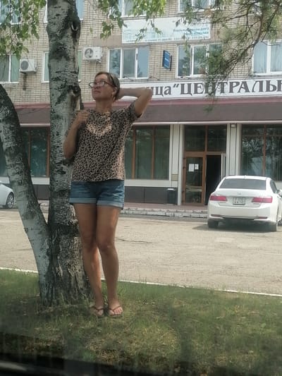 A woman in her late 20s or early 30s stands next to a tree, leaning against it with her right hand behind her head and her left hand on her hip. She is wearing a leopard-print t-shirt and denim shorts. The woman is wearing glasses and flip-flops. She is looking upwards and to her right. The background shows a building with large windows and a sign that reads "ЦЕНТРАЛЬНАЯ" (Centralnaya) in large, white letters. A white car is parked in front of the building, and another white car is partially visible on the left side of the frame. The scene appears to be a street in the city of Lesozavodsk, Russia, with a paved area in front of the building and a grassy area in the foreground. The time of day appears to be daytime.
