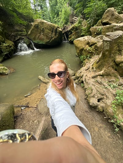In Waldbillig, Luxembourg, a woman with blonde hair, wearing sunglasses and a white knitted sweater, takes a selfie near a body of water. The water is a murky green and flows gently, with small cascades visible in the background. Large, moss-covered rocks are scattered around the water's edge, and tree roots snake across the ground. The environment is lush and green, with dense foliage in the background and sunlight filtering through the trees. A wooden staircase is visible in the distance, suggesting a well-maintained park or hiking area. The woman's expression is joyful and engaged with the camera.