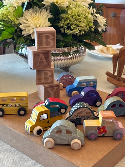 A still-life photograph on a table presents a collection of objects, including wooden blocks spelling "BABY," a bouquet of white and green flowers, a silver container, and a series of colorful wooden toy cars and a school bus. The backdrop is a light-colored tablecloth and a wall. The specific location in La Molina, Peru, cannot be determined from the image alone.